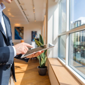 A person in formal attire is using a tablet while standing near a large window. The interior is bright and modern, featuring wooden flooring and potted plants. Artwork is displayed on the walls, adding a touch of character to the space.