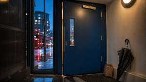 A deep-blue lobby fire door closes beside a wet black umbrella and grocery bag during fire door checks.